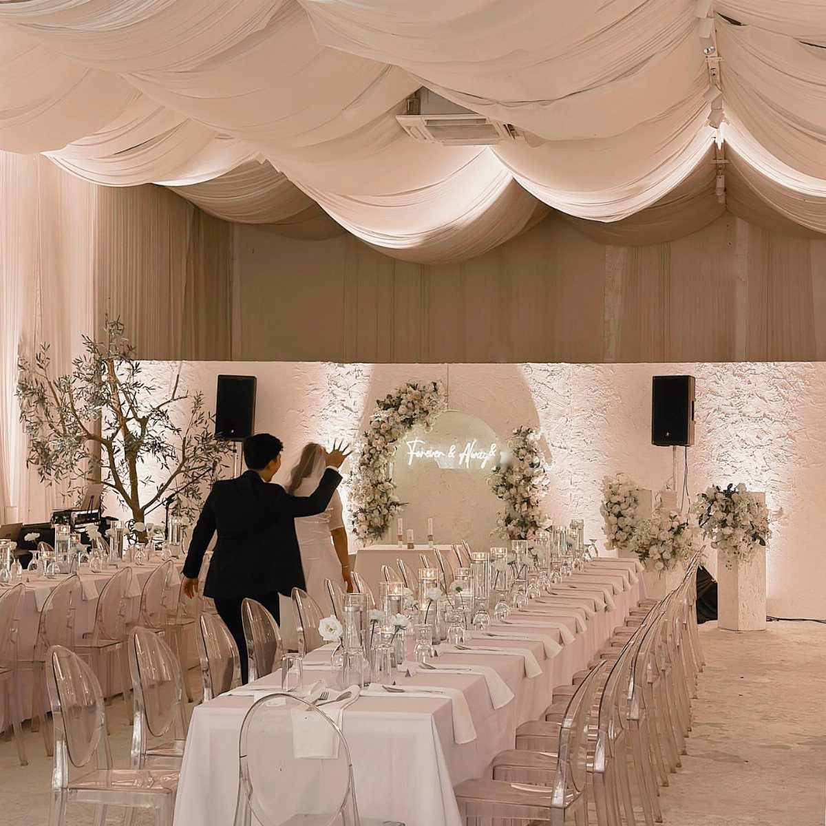White wedding reception with clear acrylic chairs, draped ceiling, and floral arrangements.