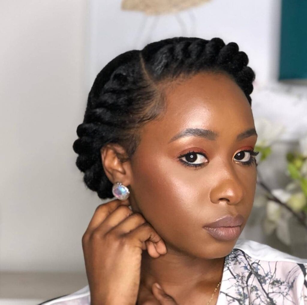 Black woman with a twisted crown braid hairstyle and warm-toned makeup.