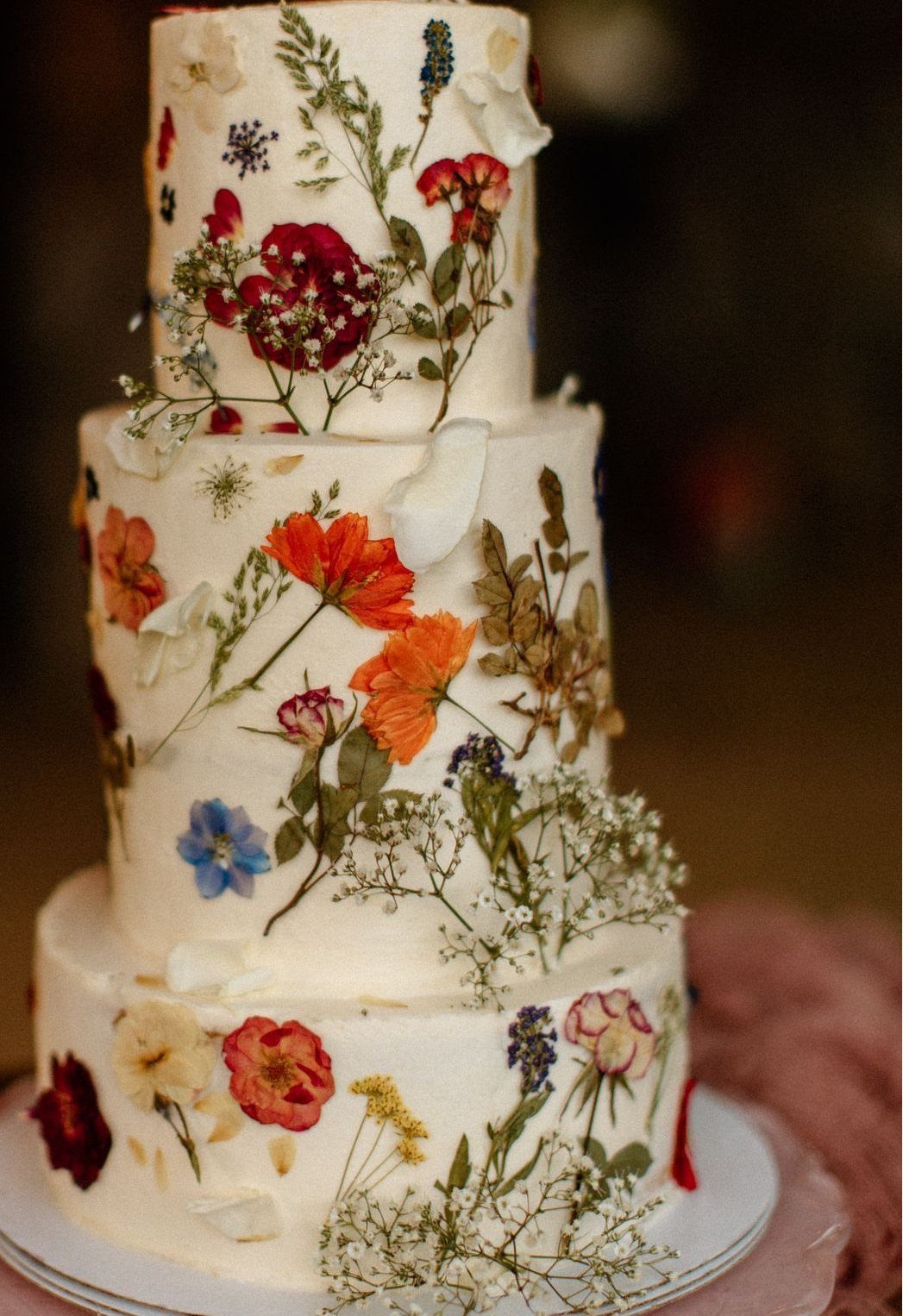 Three-tiered white wedding cake decorated with colorful pressed wildflowers and greenery.