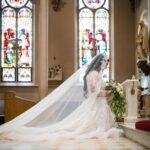 Bride in white lace gown and veil kneeling in a church with stained glass.