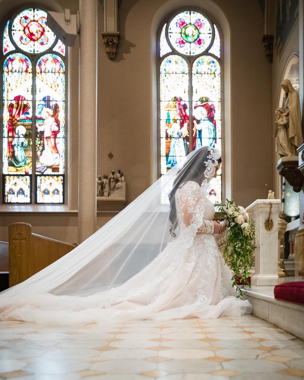 Bride in white lace gown and veil kneeling in a church with stained glass.