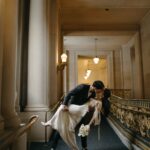 Bride and groom kissing in a grand hallway with stone columns and gold railings.
