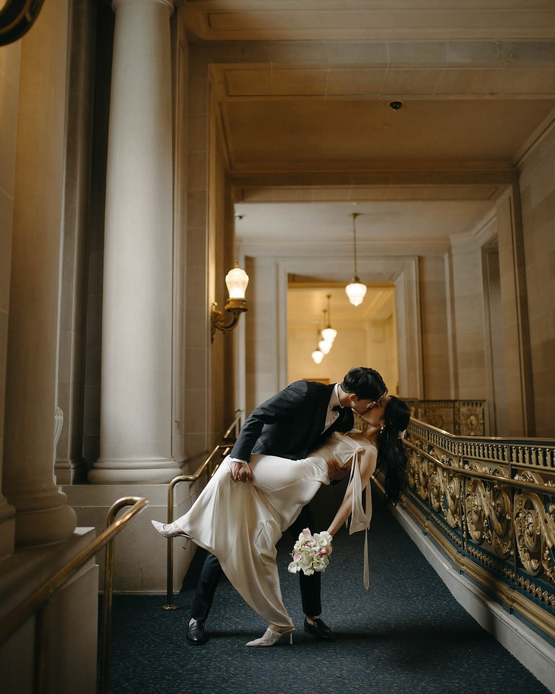 Bride and groom kissing in a grand hallway with stone columns and gold railings.