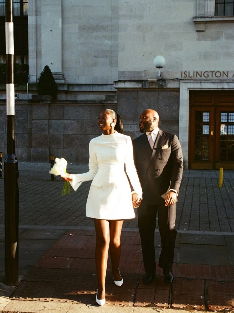 Couple in wedding attire walking outside a building, woman in short white dress.