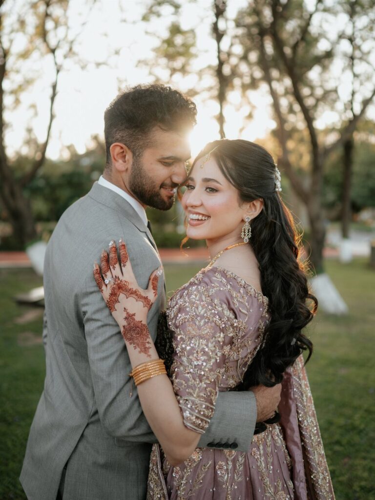 Smiling couple in wedding attire posing closely outdoors with sunlight filtering through trees.