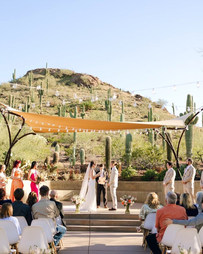 Wedding ceremony in a desert landscape with saguaro cacti and a tan fabric canopy.