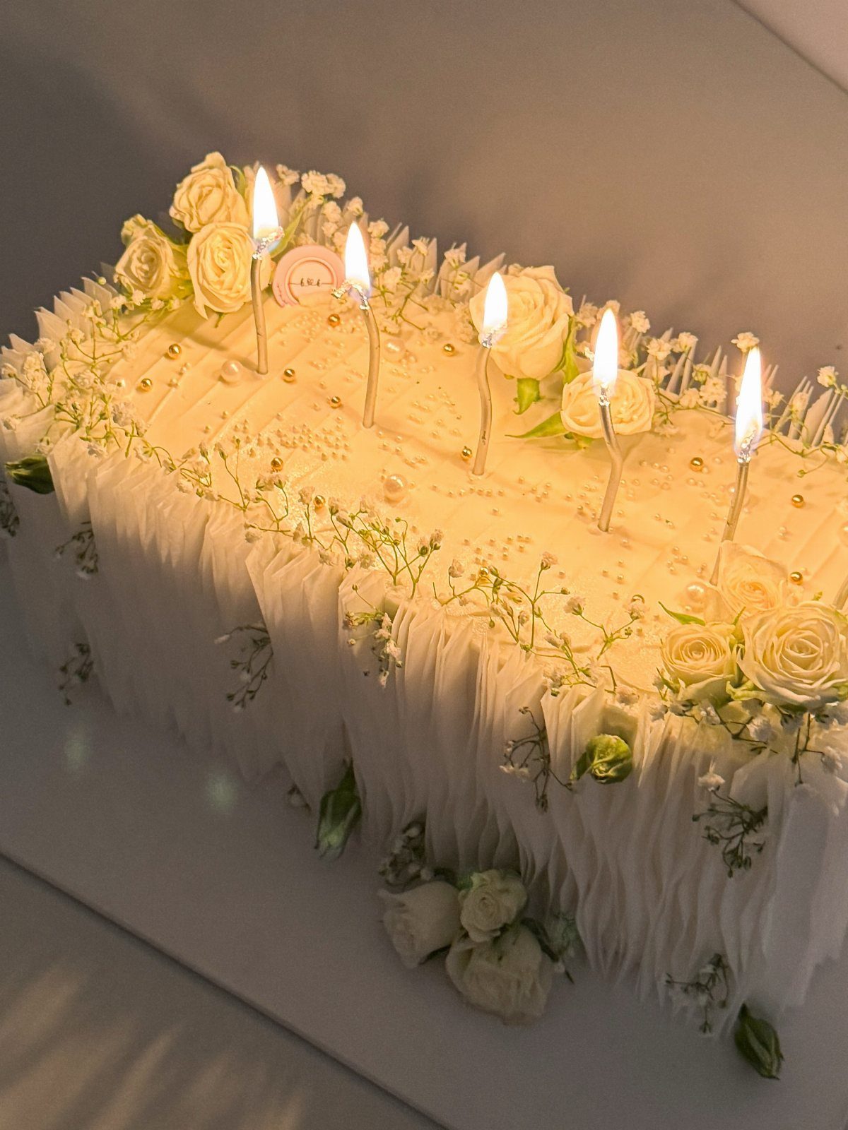 Flat white rectangular wedding cake decorated with white roses and five lit candles.