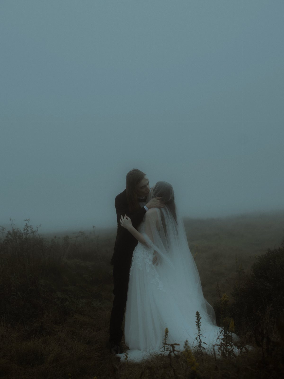 Bride and groom embracing in a dark, foggy field with a moody atmosphere.