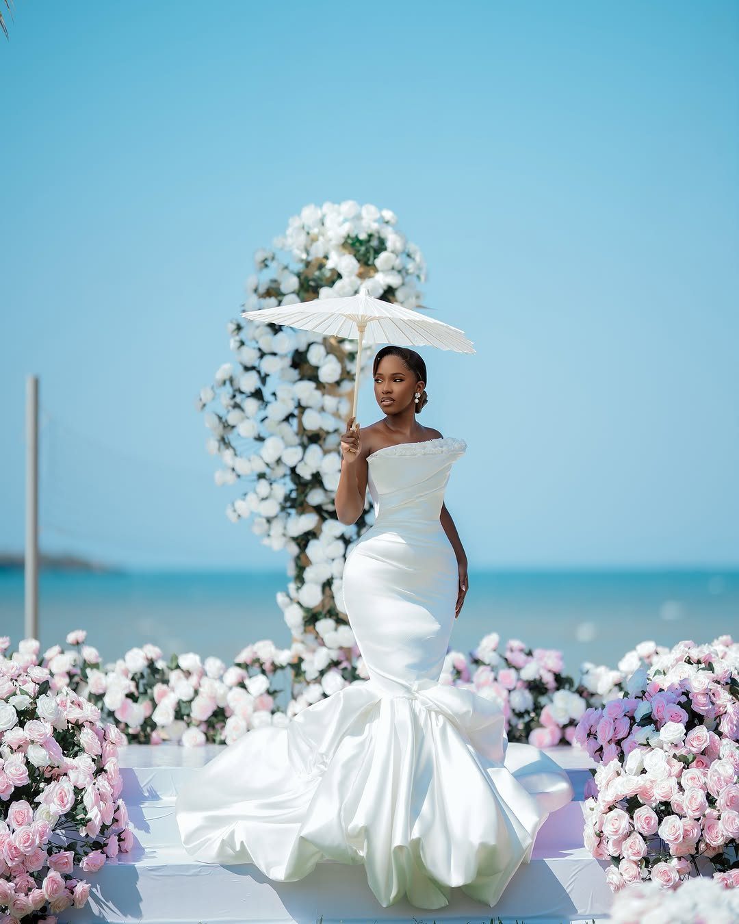 Woman in white off-the-shoulder mermaid wedding dress holding a parasol by the ocean.