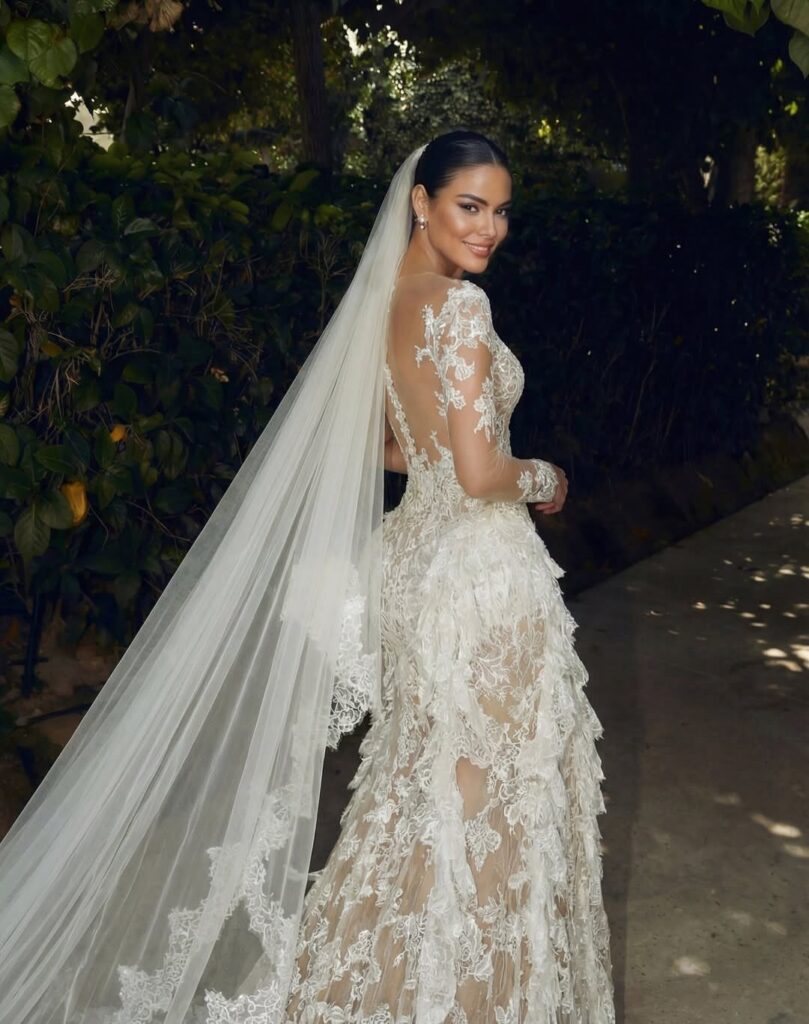 Woman in a long white lace wedding dress and veil posing outdoors.