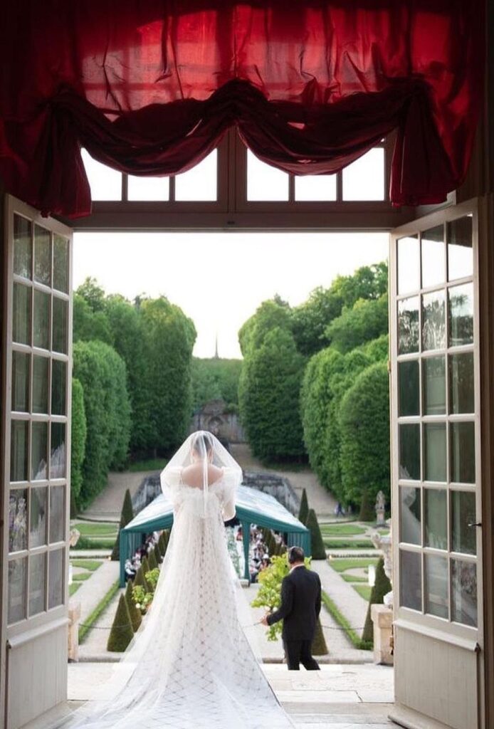 Bride in white gown looking out from a doorway onto a formal garden.