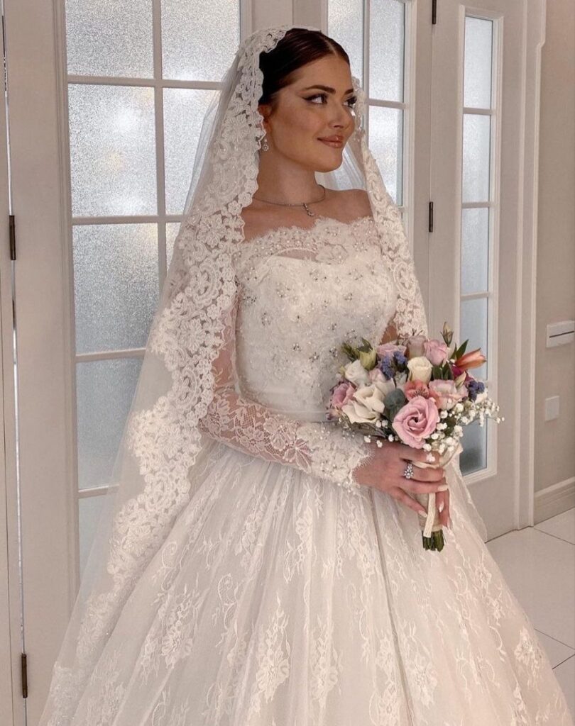 Bride in a white lace wedding dress and mantilla veil holding a rose bouquet.