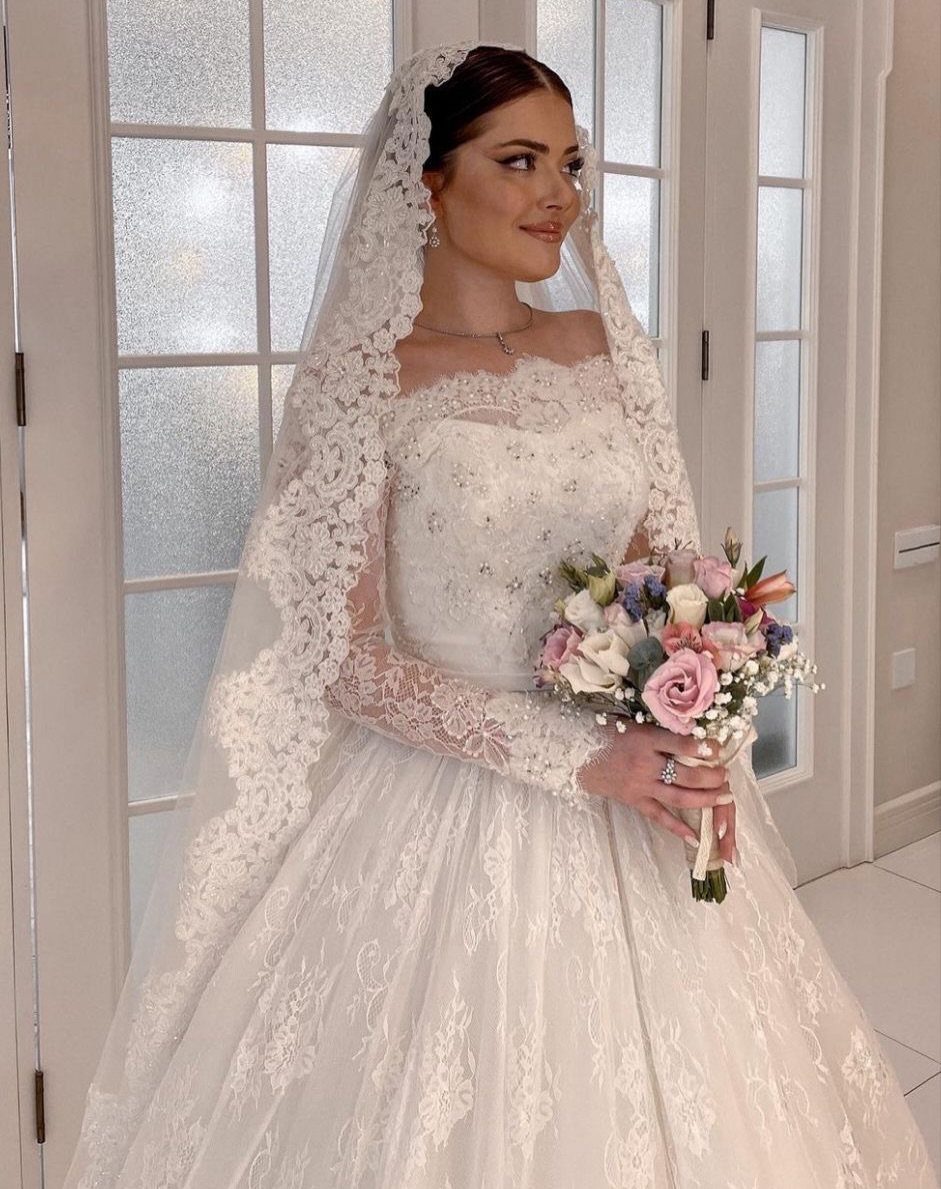 Bride in a white lace wedding dress and mantilla veil holding a rose bouquet.