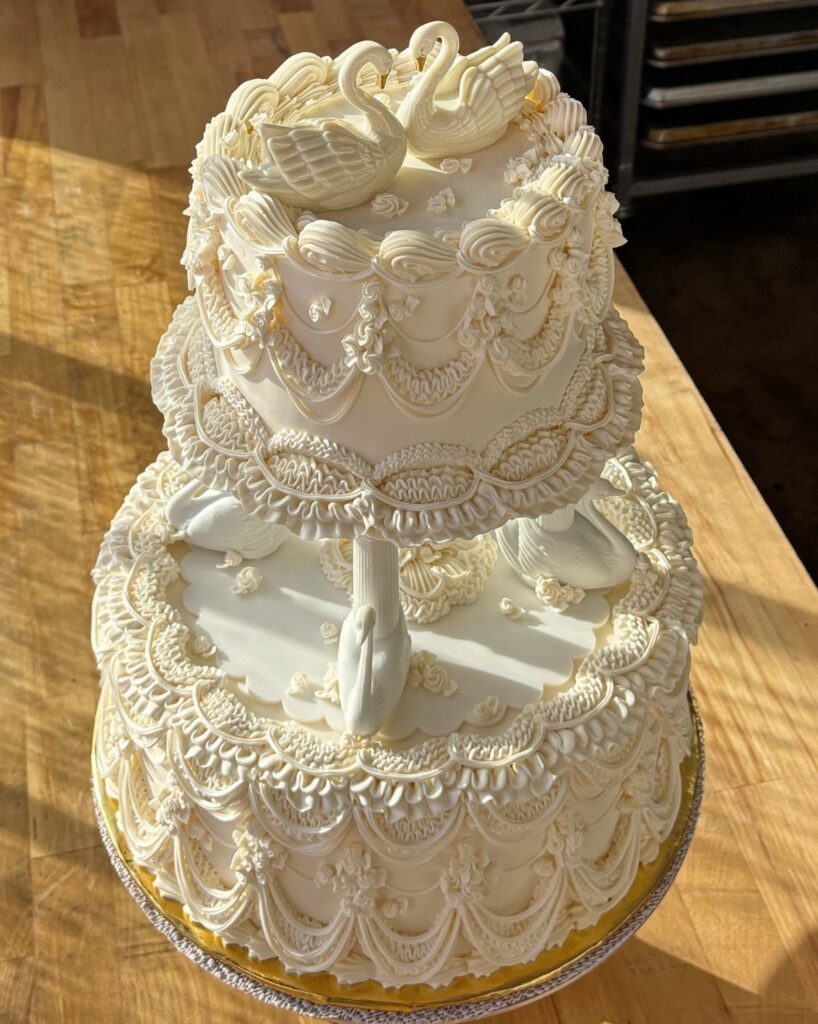 Three-tiered white wedding cake with ornate frosting swags and sculpted swans on a table.