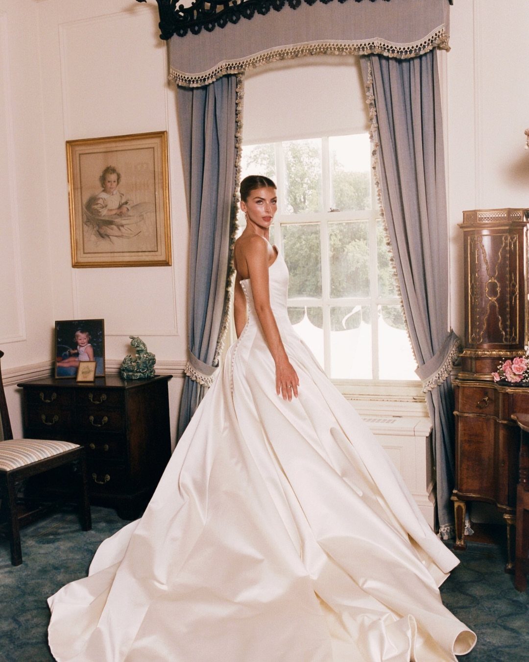 Woman in a voluminous white strapless wedding gown posing by a window with blue curtains.