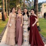 Four women wearing formal floor-length gowns posing outdoors among palm trees at dusk.