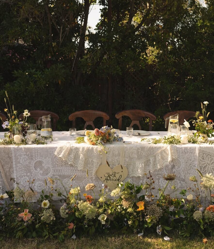 Low floral centerpieces and greenery on a white lace wedding table outdoors.