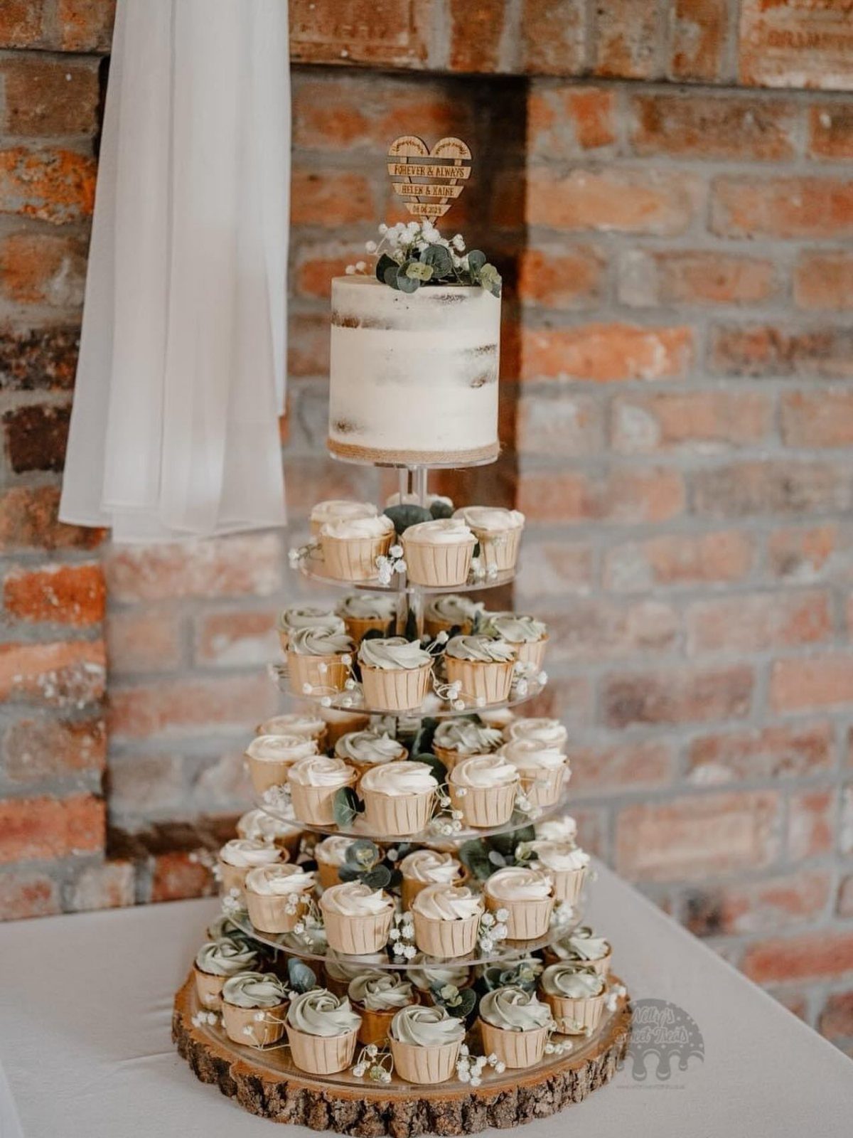 Rustic wedding cake and cupcake tower on a wood slice against brick wall.