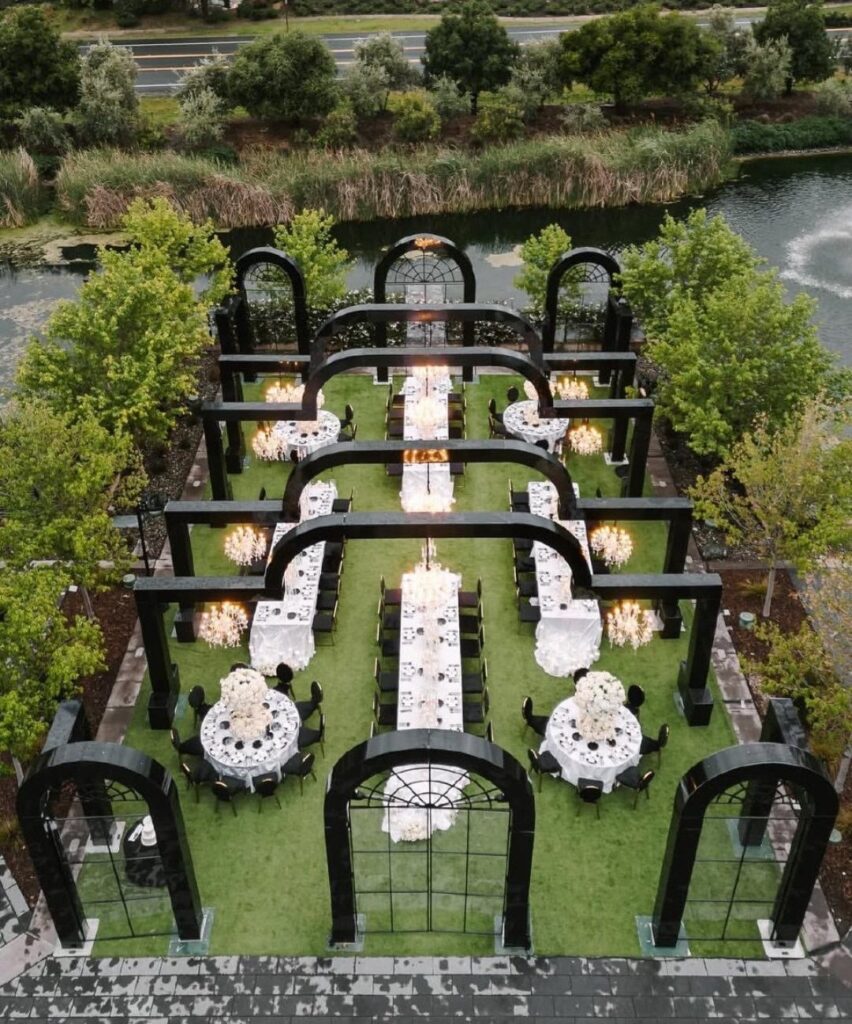 Outdoor wedding reception setup with white tables and black arches on a green lawn.