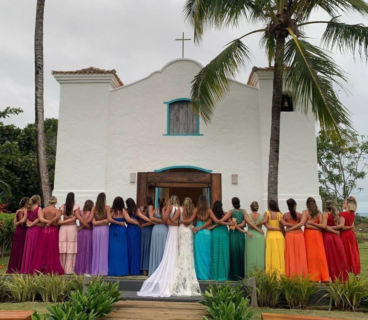 Bridesmaids in rainbow colored dresses standing before a white church with palm trees.