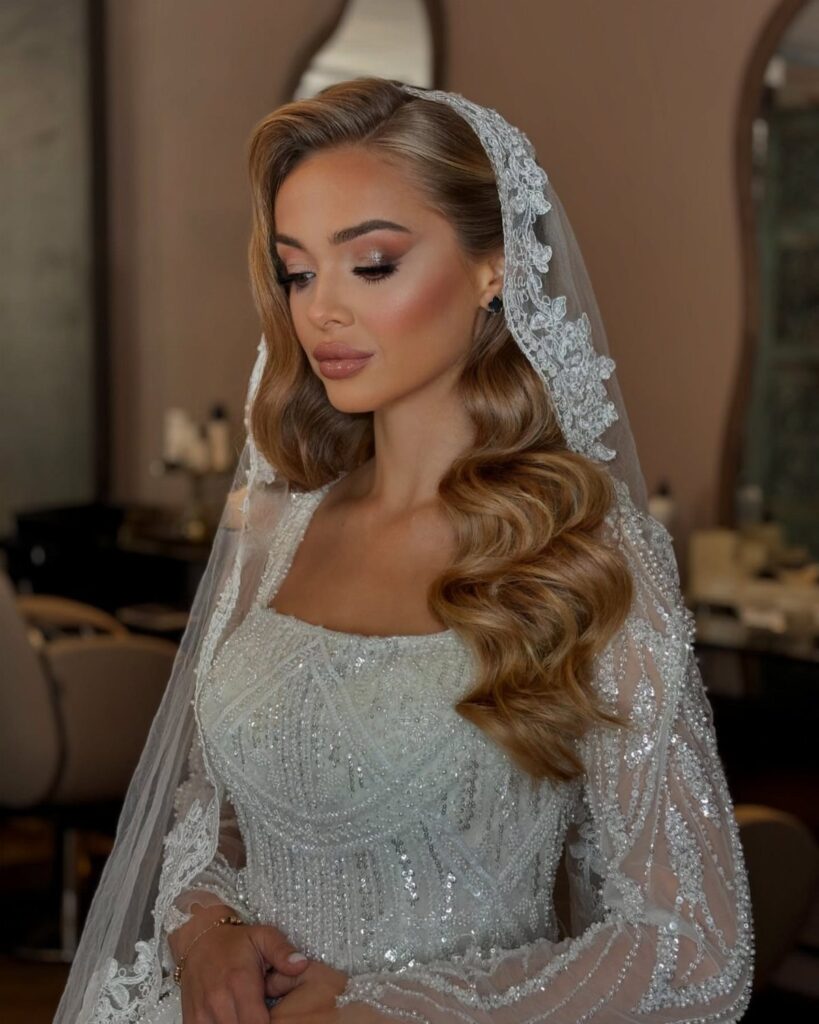 Woman with wavy blonde hair wearing a white bridal gown and lace veil.