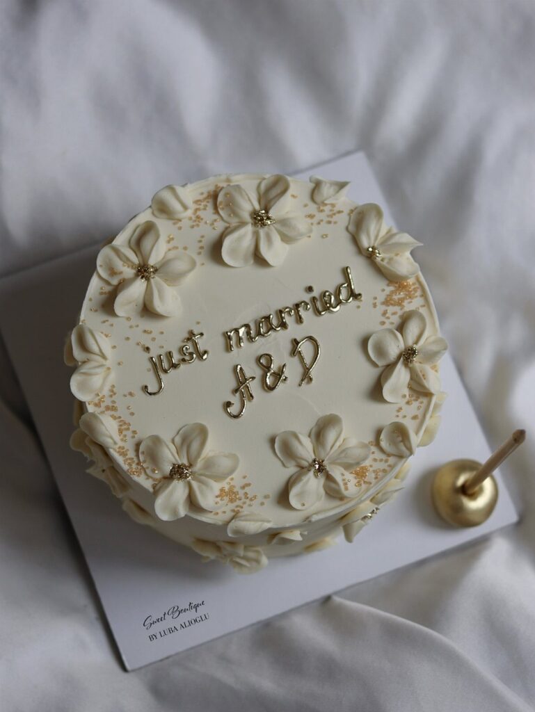 Small single-tier white wedding cake with gold lettering and frosting flowers on white background