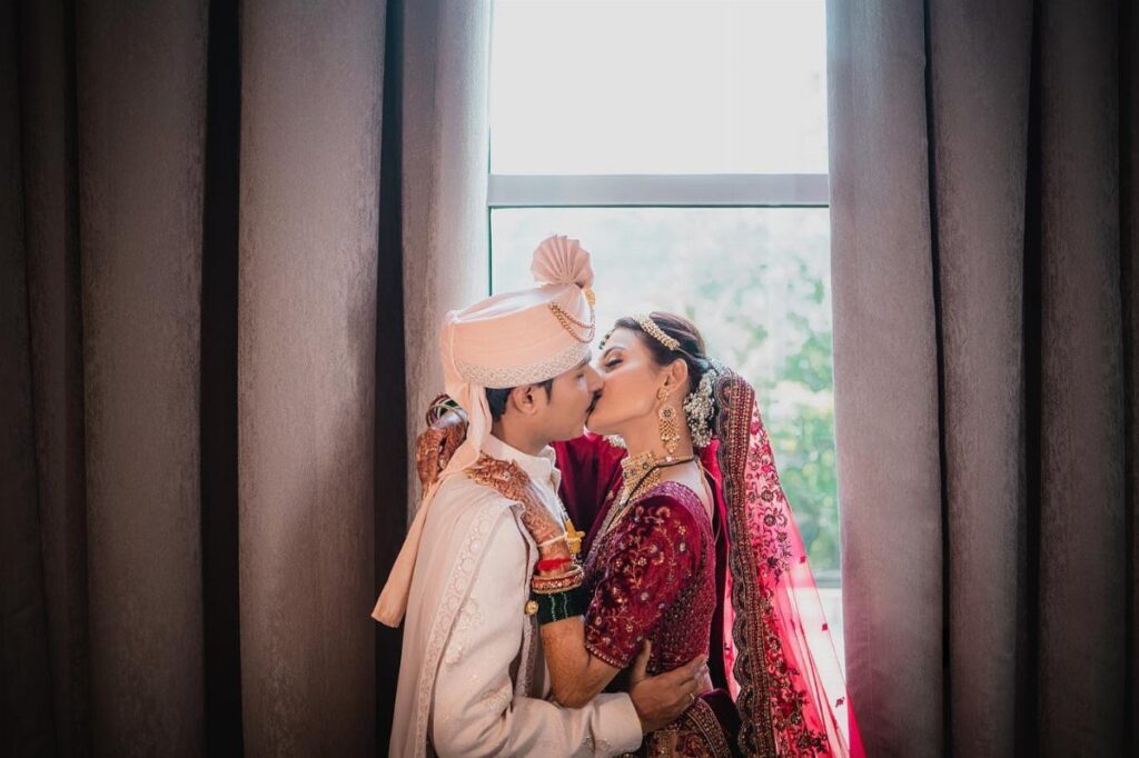 Indian bride and groom in traditional wedding attire kissing by a bright window