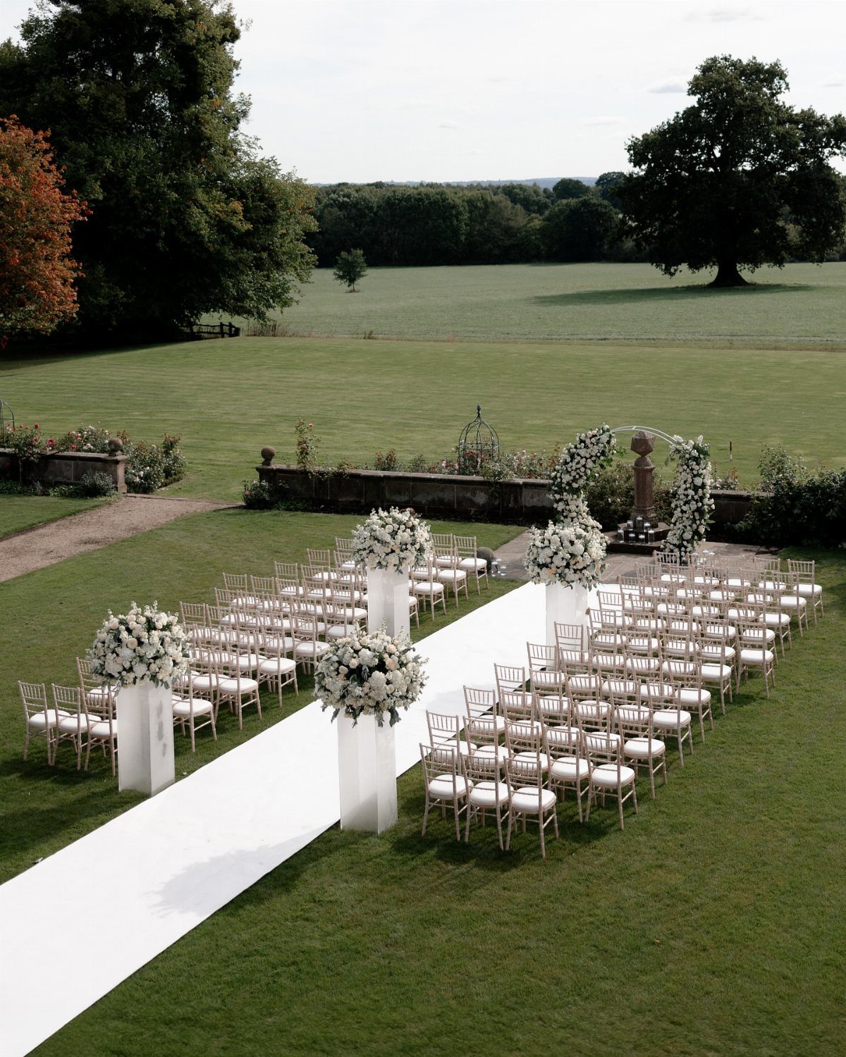 Outdoor wedding ceremony setup with white chairs, floral arrangements, and a white aisle runner.